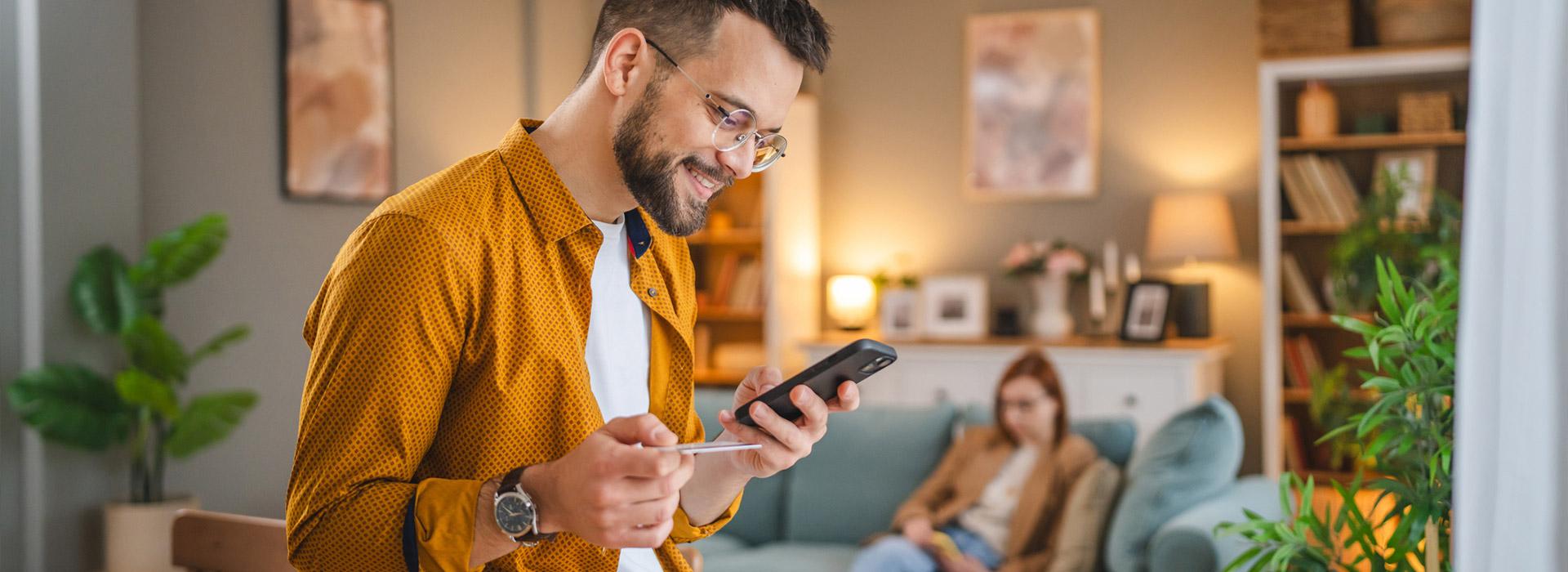 Man in orange shirt with credit card in one hand and cell phone in the other. Woman sitting down on sofa in the distant background.